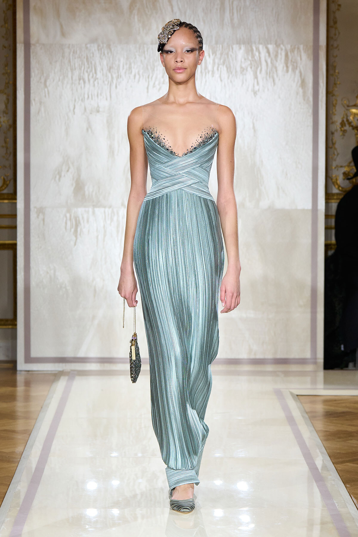 Model on a runway wearing a pale blue-green pleated gown with a beaded neckline and a matching headpiece, carrying a small clutch.