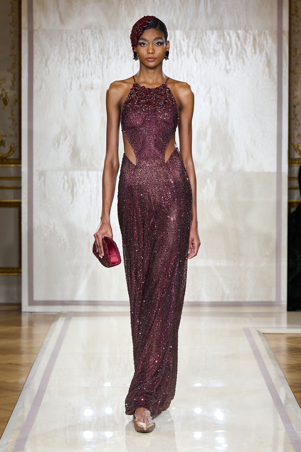 Model on a runway wearing a burgundy sequined halter gown with side cutouts and matching burgundy clutch; ornate backdrop behind her.