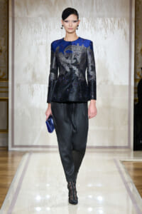 Runway model wearing a blue and black sequined jacket with lace embroidery, paired with black satin pants, black boots, and a blue clutch.