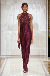 Model walks the runway in a burgundy beaded halter gown with intricate lace bodice and vertical pleated skirt, holding a matching clutch.