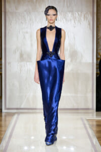 Model on a runway wearing a glossy deep blue gown with a plunging neckline and black jeweled accessories, hair pinned with a floral headpiece.