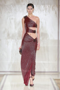 Model on the runway wearing a burgundy glitter one-shoulder gown with cutouts and jeweled floral accents