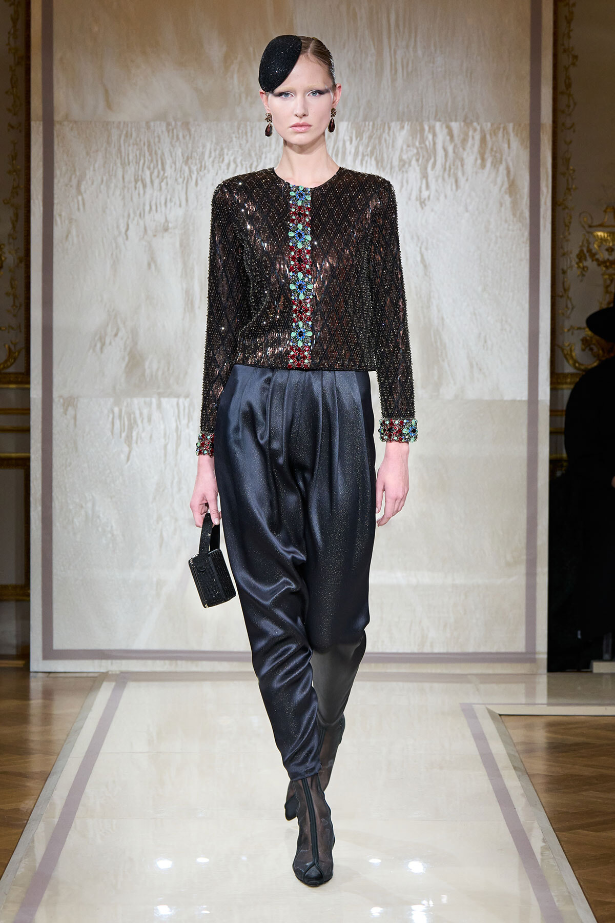 Model on the runway wearing a black sequined jacket with a colorful beaded center stripe and cuffs, paired with dark satin trousers and a small black handbag.