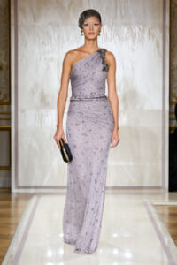 Female model walks a lavender one-shoulder gown with beaded embellishments, holding a black clutch on a glossy runway.