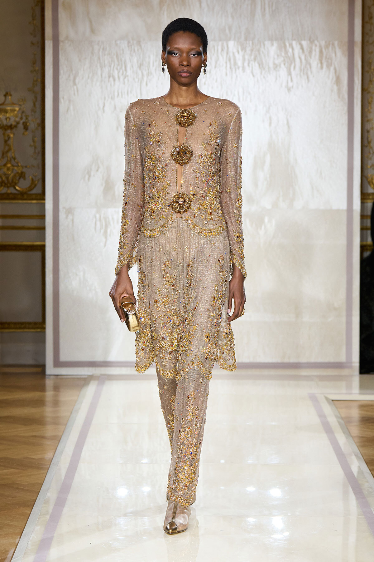 Model wearing a sheer, gold-embellished gown with long sleeves and circular jewel accents walking a runway, holding a small gold clutch.