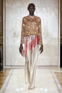 Model walks the runway in a gold beaded, sequined jacket with pink accents paired with cream brocade wide-leg pants, clutch in hand.