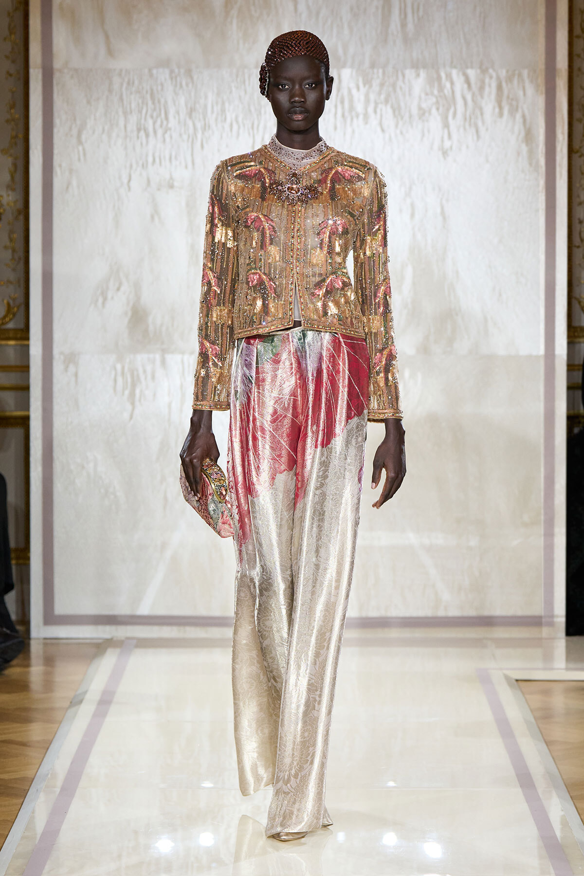 Model walks the runway in a gold beaded, sequined jacket with pink accents paired with cream brocade wide-leg pants, clutch in hand.