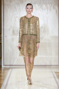 Model walking a runway in an opulent gold beaded lace dress with green accents and matching heels.
