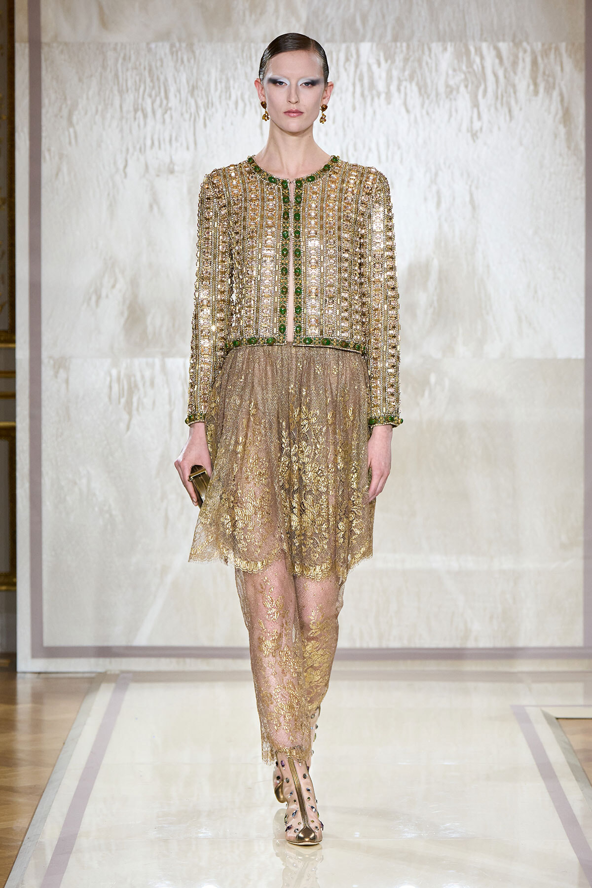 Model walking a runway in an opulent gold beaded lace dress with green accents and matching heels.