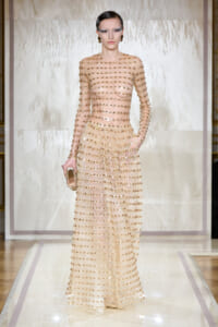 Model on a runway wearing a sheer, full-length nude gown covered in gold studs, holding a small clutch.