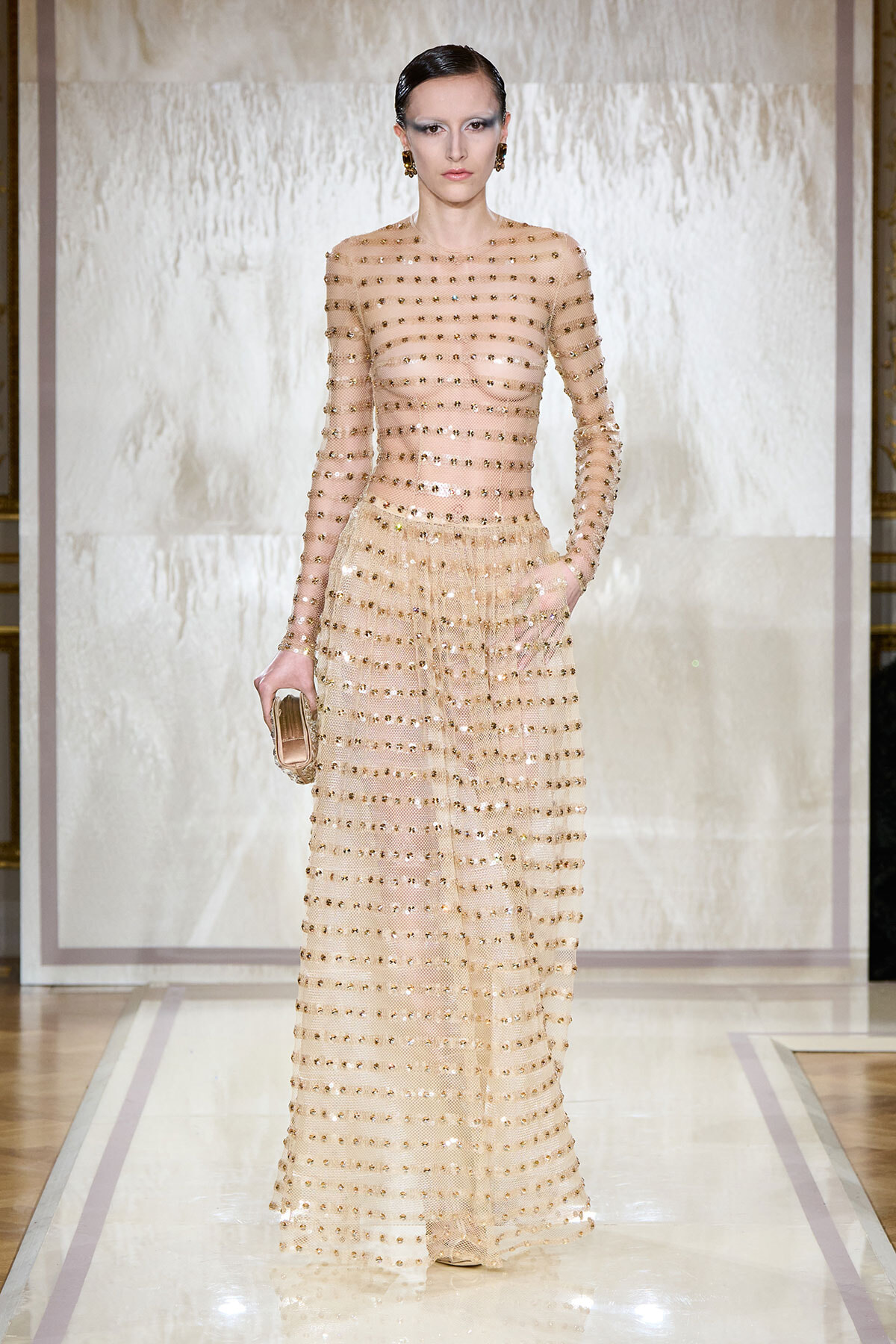 Model on a runway wearing a sheer, full-length nude gown covered in gold studs, holding a small clutch.
