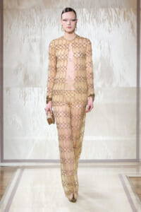 Model on runway wearing a sheer gold chain-link gown over a nude base, with matching jewelry and a small clutch underline the look.