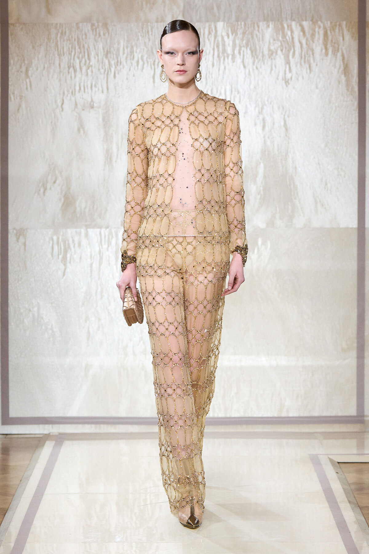 Model on runway wearing a sheer gold chain-link gown over a nude base, with matching jewelry and a small clutch underline the look.