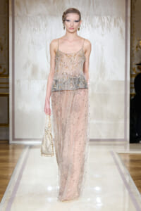 Model walking a runway in a sheer, beaded gown with gold floral embroidery and a matching headpiece; carrying a small beaded handbag.