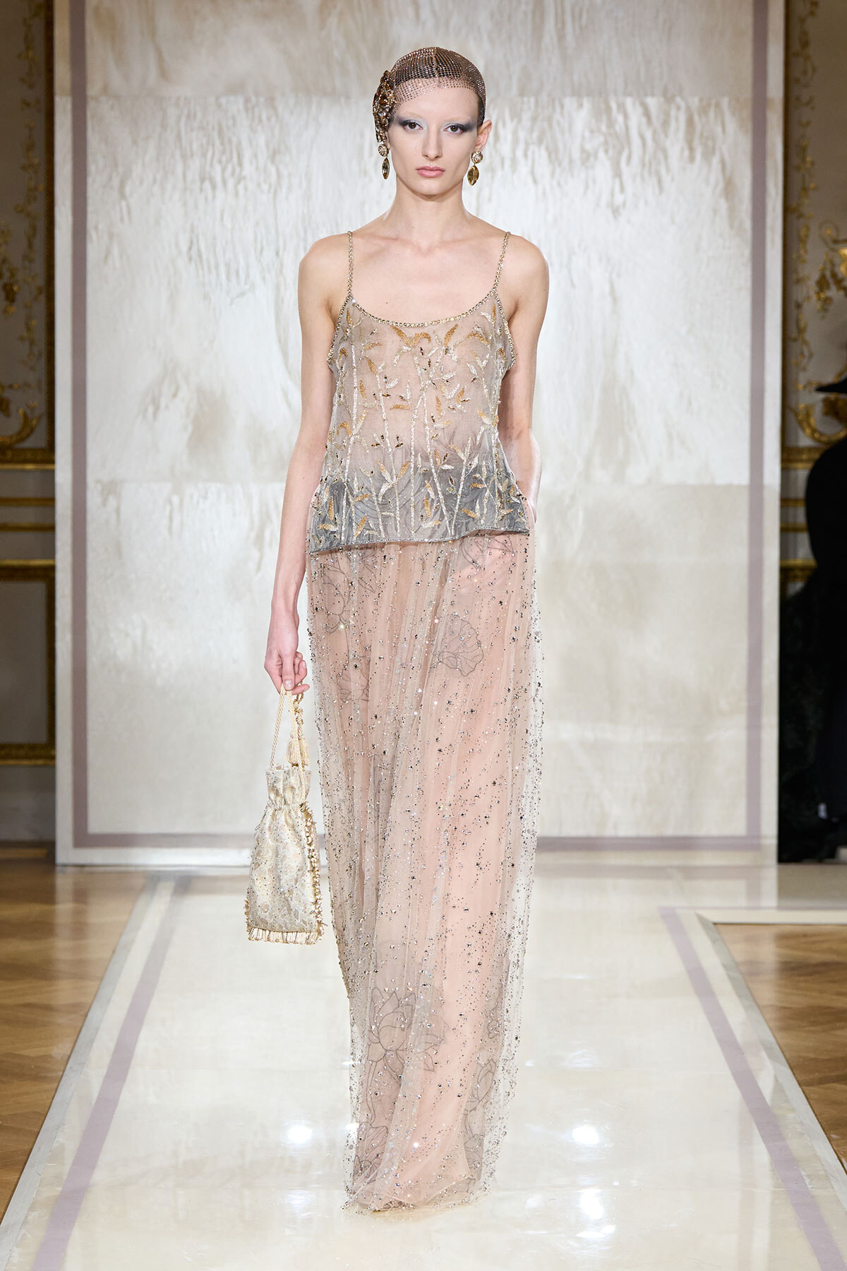 Model walking a runway in a sheer, beaded gown with gold floral embroidery and a matching headpiece; carrying a small beaded handbag.