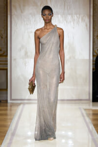 Model on a runway wearing a silver one-shoulder glitter gown, holding a gold clutch.