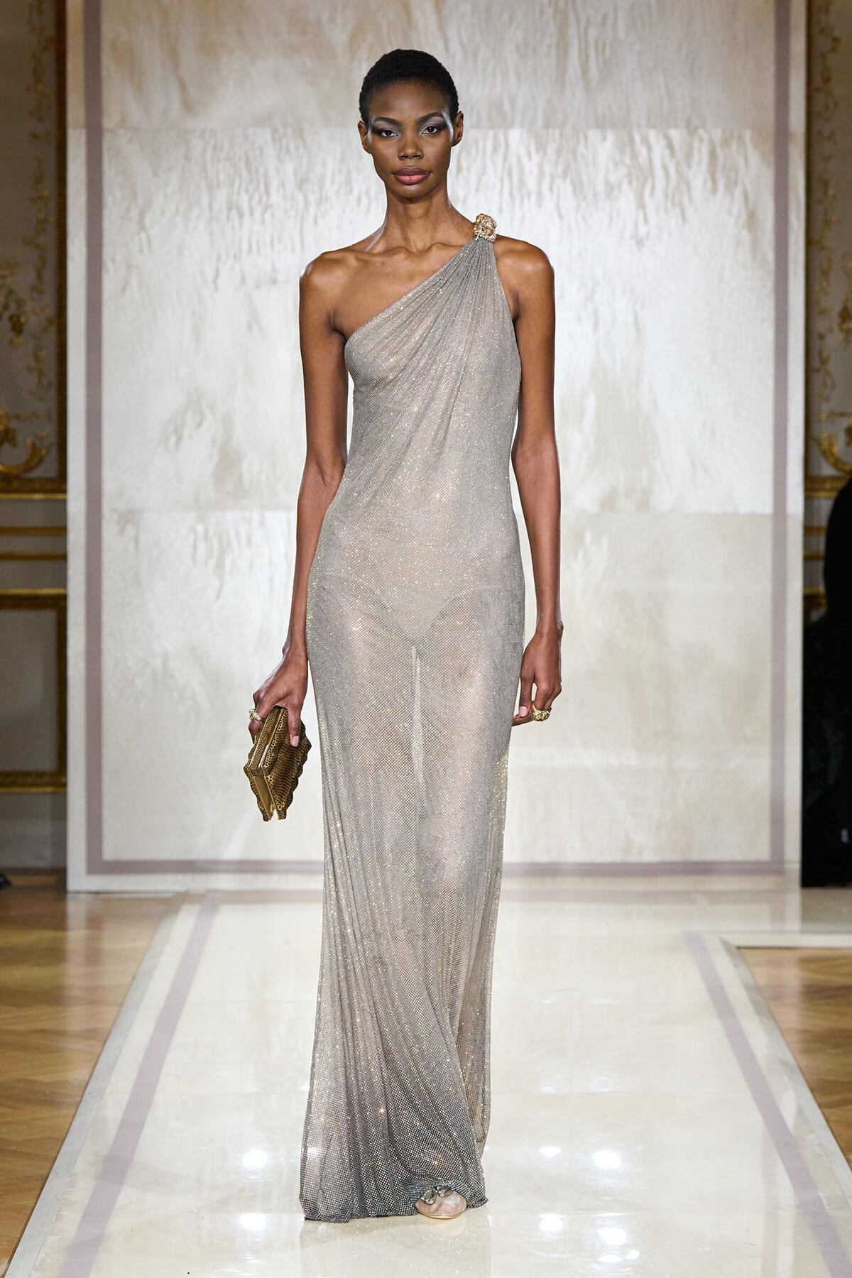 Model on a runway wearing a silver one-shoulder glitter gown, holding a gold clutch.