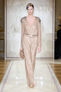 Model walking the runway in a champagne gold sequined wrap jumpsuit with jeweled shoulders, belt, headscarf, and matching clutch.