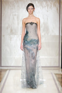 Model on a runway wearing a strapless silver-gray gown with a sheer, beaded overlay and blue-green embellishments at the hips and bodice.