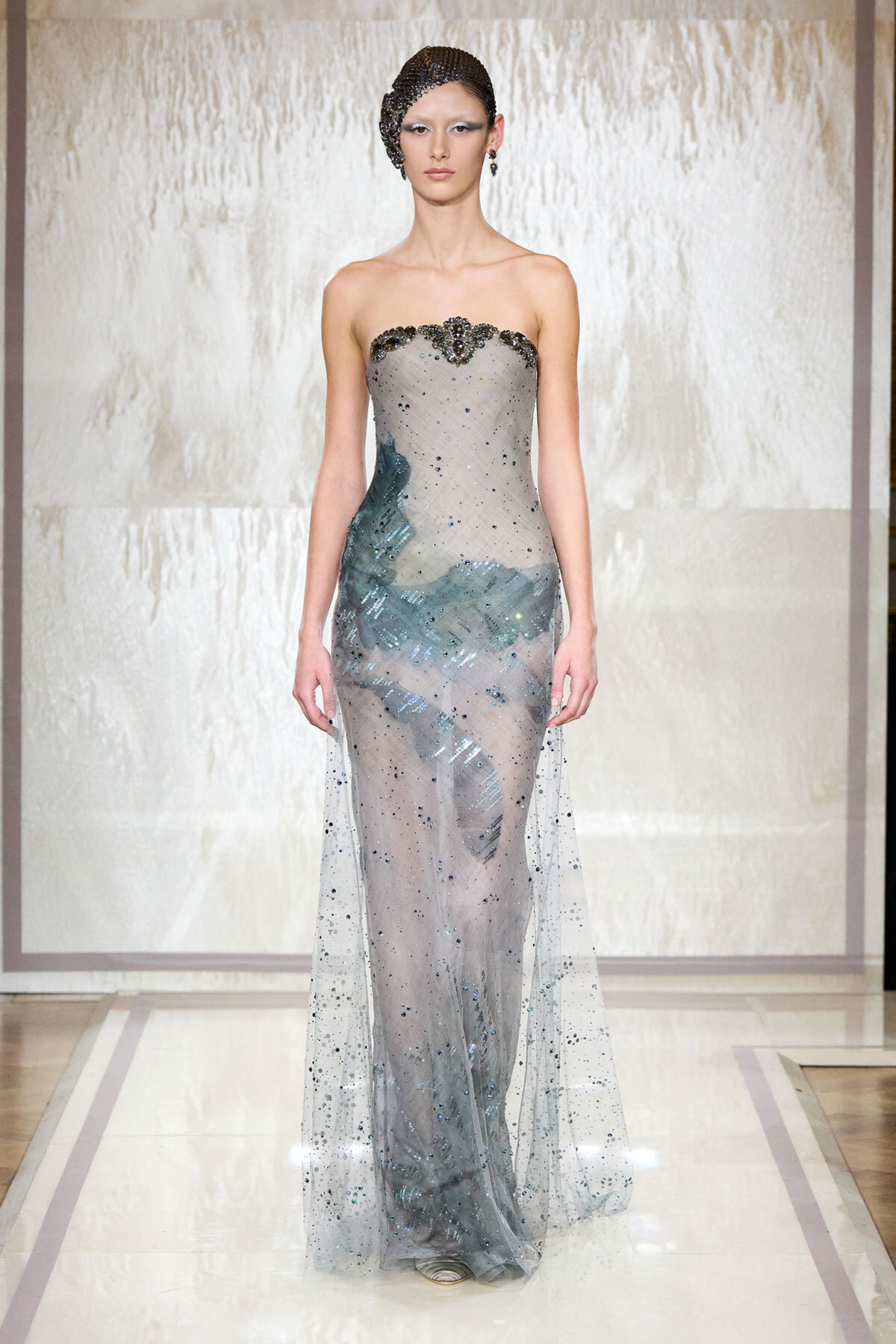 Model on a runway wearing a strapless silver-gray gown with a sheer, beaded overlay and blue-green embellishments at the hips and bodice.