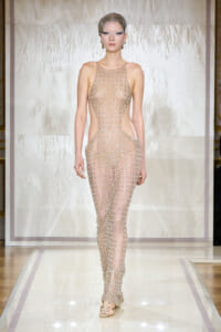 Model walking a runway in a nude, beaded cutout jumpsuit with matching headpiece and earrings on a glossy catwalk.