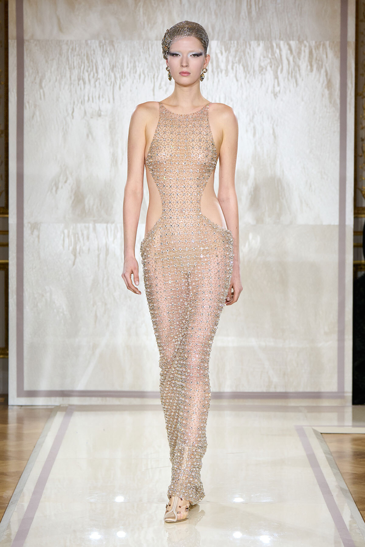 Model walking a runway in a nude, beaded cutout jumpsuit with matching headpiece and earrings on a glossy catwalk.