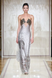 Model on a runway wearing a strapless silver gown with a beaded metallic bodice and a sheer, embroidered skirt featuring palm frond design.