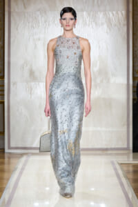 Model walking runway in a floor-length silver sequin gown with a high neck and bead detail, carrying a small metallic box purse.