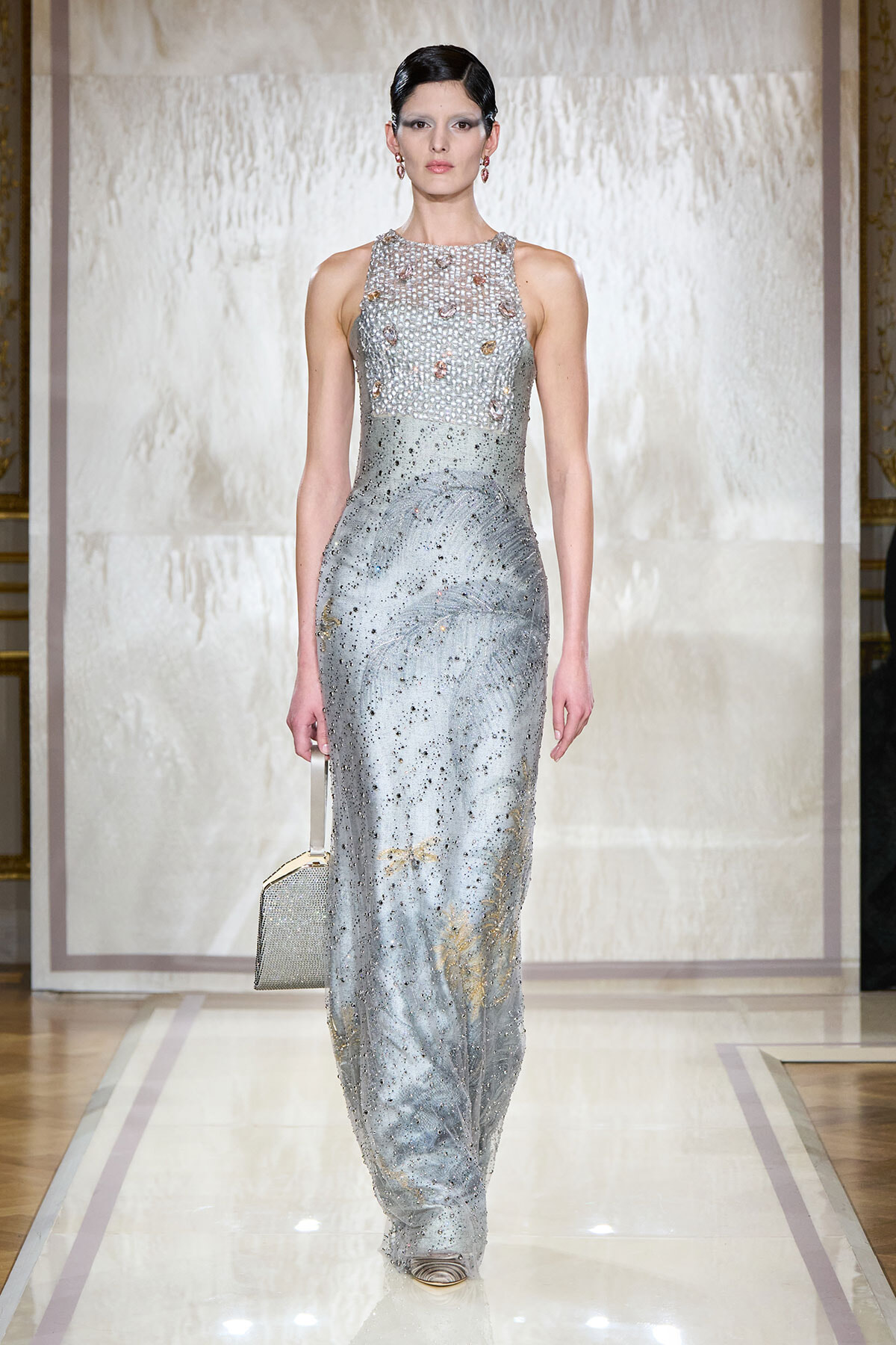 Model walking runway in a floor-length silver sequin gown with a high neck and bead detail, carrying a small metallic box purse.
