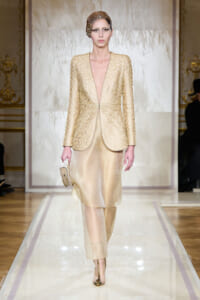 Model walking runway in a gold lace blazer and matching champagne skirt, holding a small clutch with a jeweled headpiece