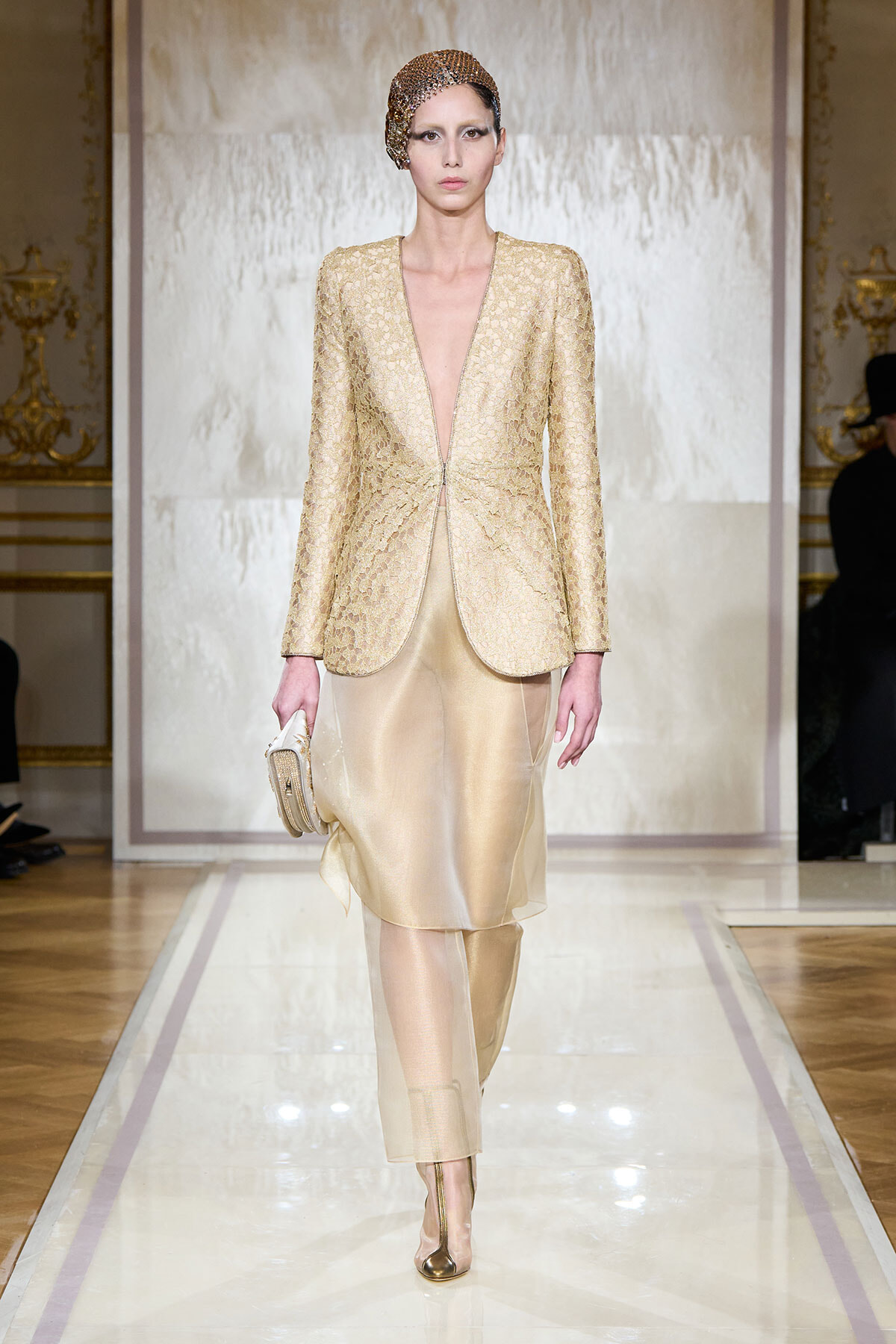 Model walking runway in a gold lace blazer and matching champagne skirt, holding a small clutch with a jeweled headpiece
