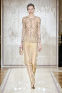 Model on a runway wearing a beaded nude sheer jacket and champagne satin pants with a matching headpiece and clutch accessory.