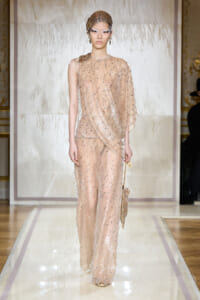 Model walking a fashion runway in a nude-pink, sequined pantsuit with a sheer draped cape and matching beaded headpiece.
