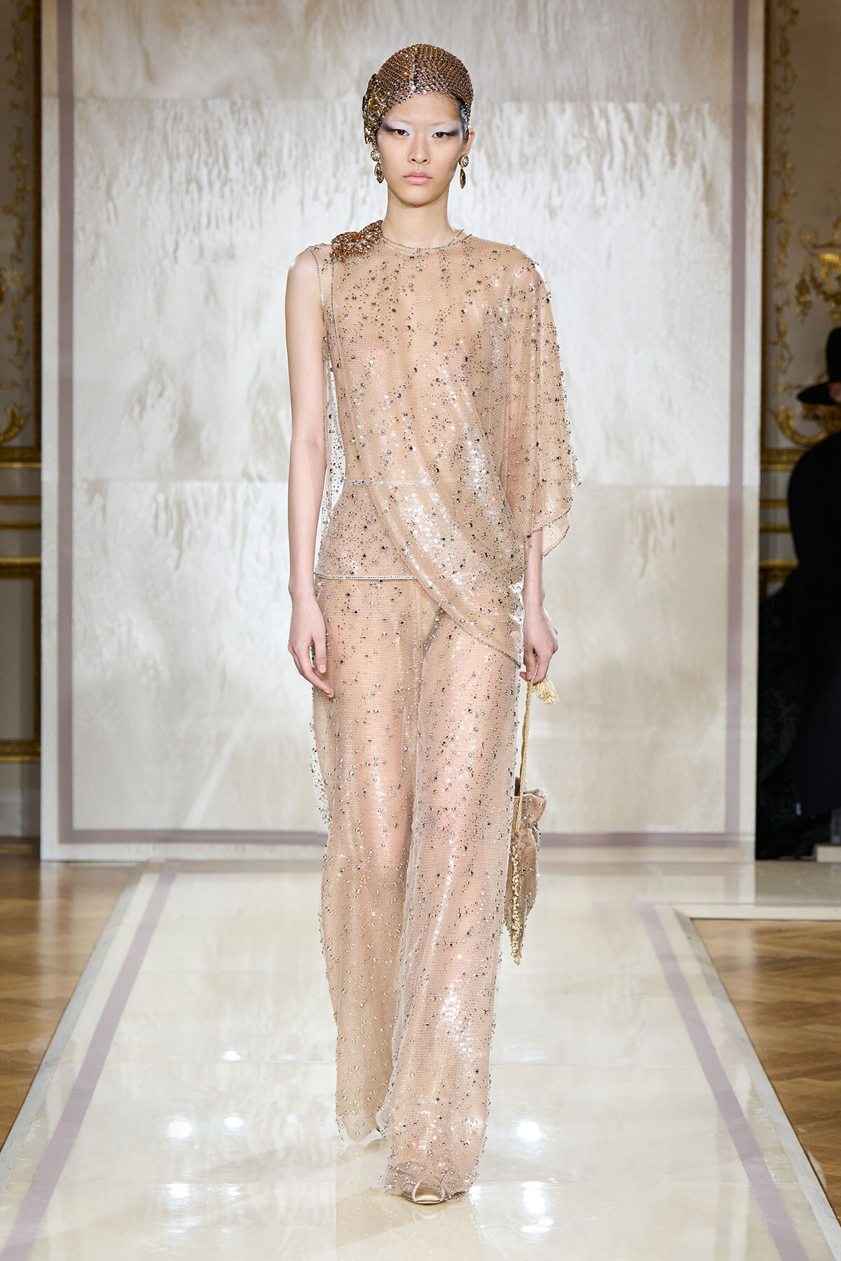 Model walking a fashion runway in a nude-pink, sequined pantsuit with a sheer draped cape and matching beaded headpiece.
