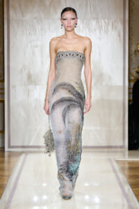 Model wearing a strapless, silver-gray gown with palm tree embroidery walking the runway on a glossy catwalk.