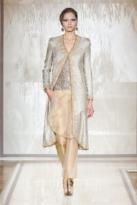 Runway model wearing a silver metallic coat with gold trim over a sequined top and beige pants, with gold accessories on a bright catwalk.