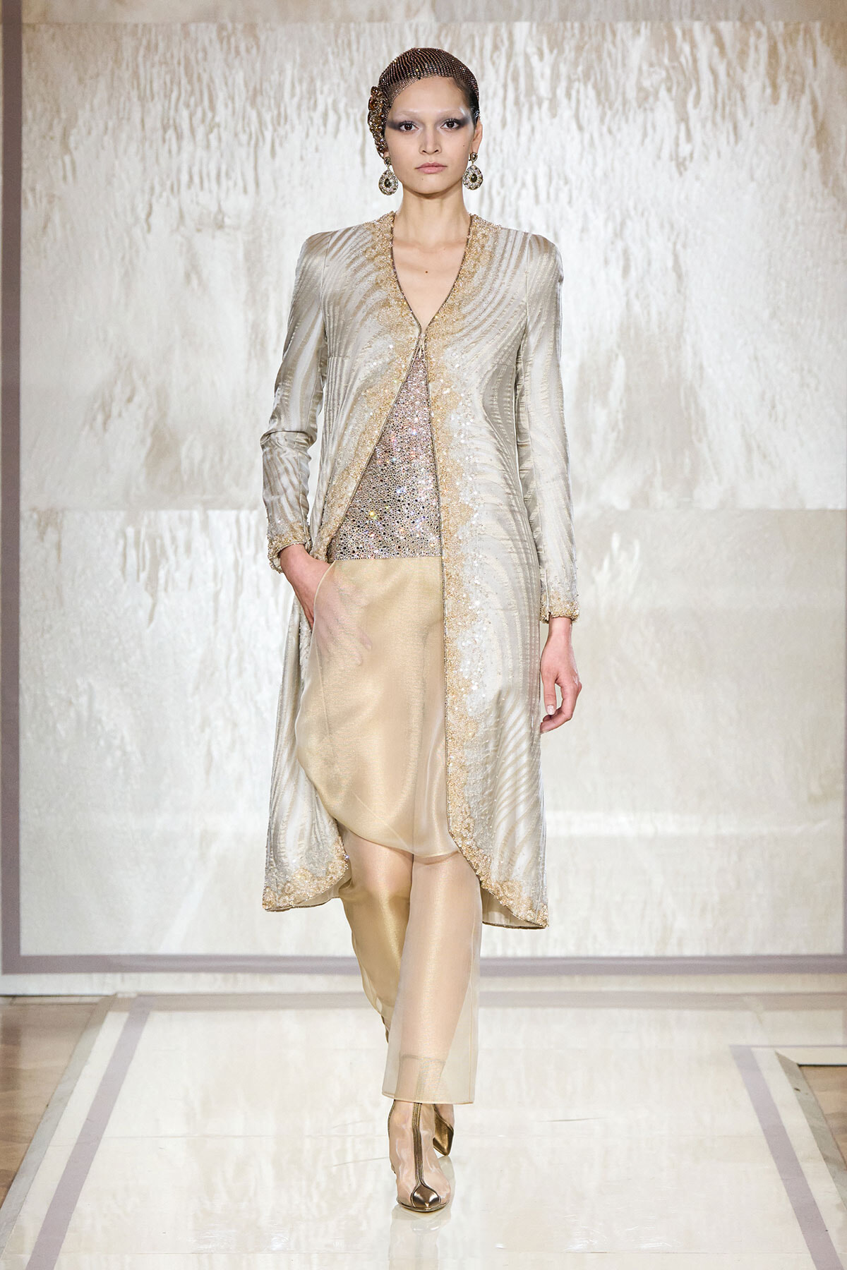 Runway model wearing a silver metallic coat with gold trim over a sequined top and beige pants, with gold accessories on a bright catwalk.