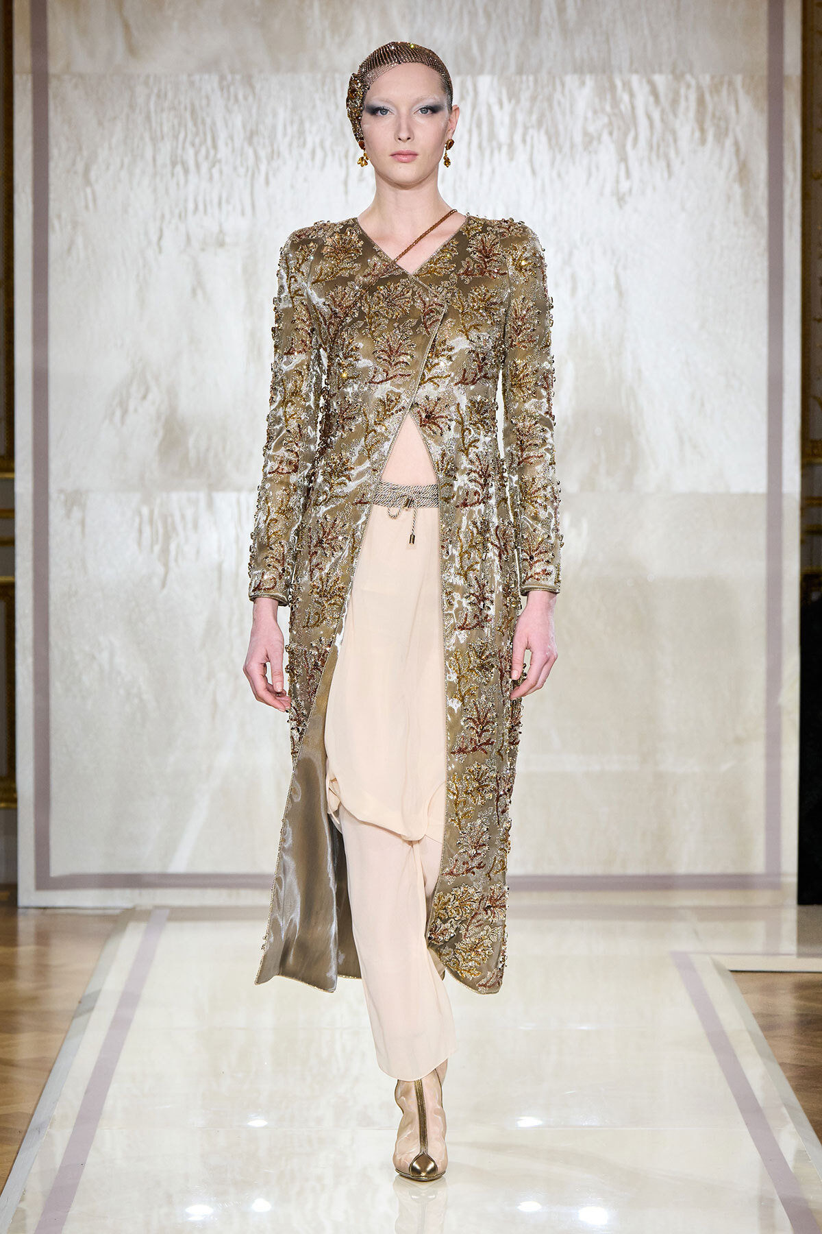 Model on a runway wearing a long gold-embellished coat with intricate floral embroidery over pale beige draped pants, gold footwear and headpiece