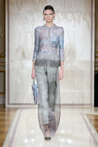 Model walks a glamorous lavender beaded jacket with sheer, wide-leg pants on a runway, holding a small patterned bag.