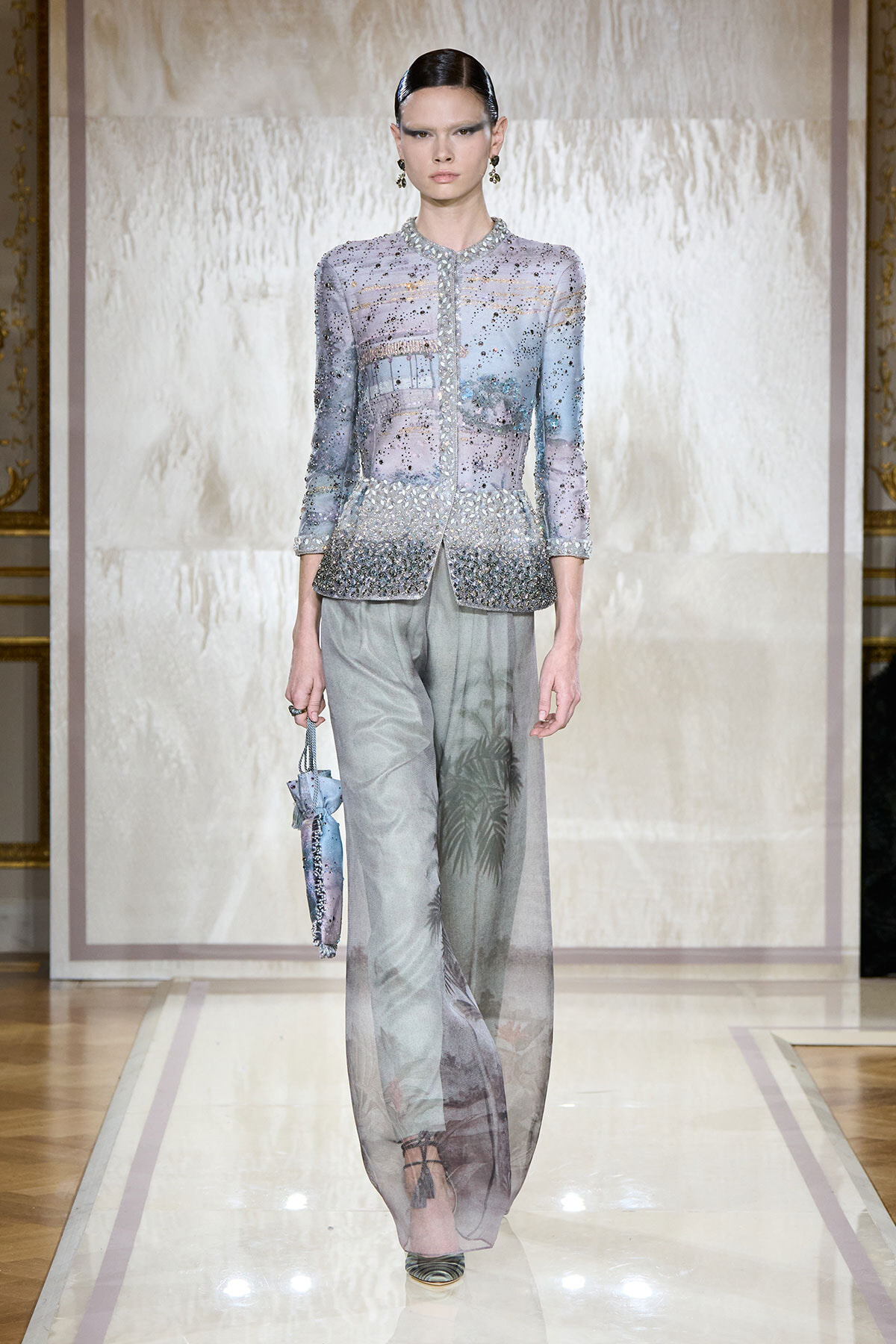 Model walks a glamorous lavender beaded jacket with sheer, wide-leg pants on a runway, holding a small patterned bag.