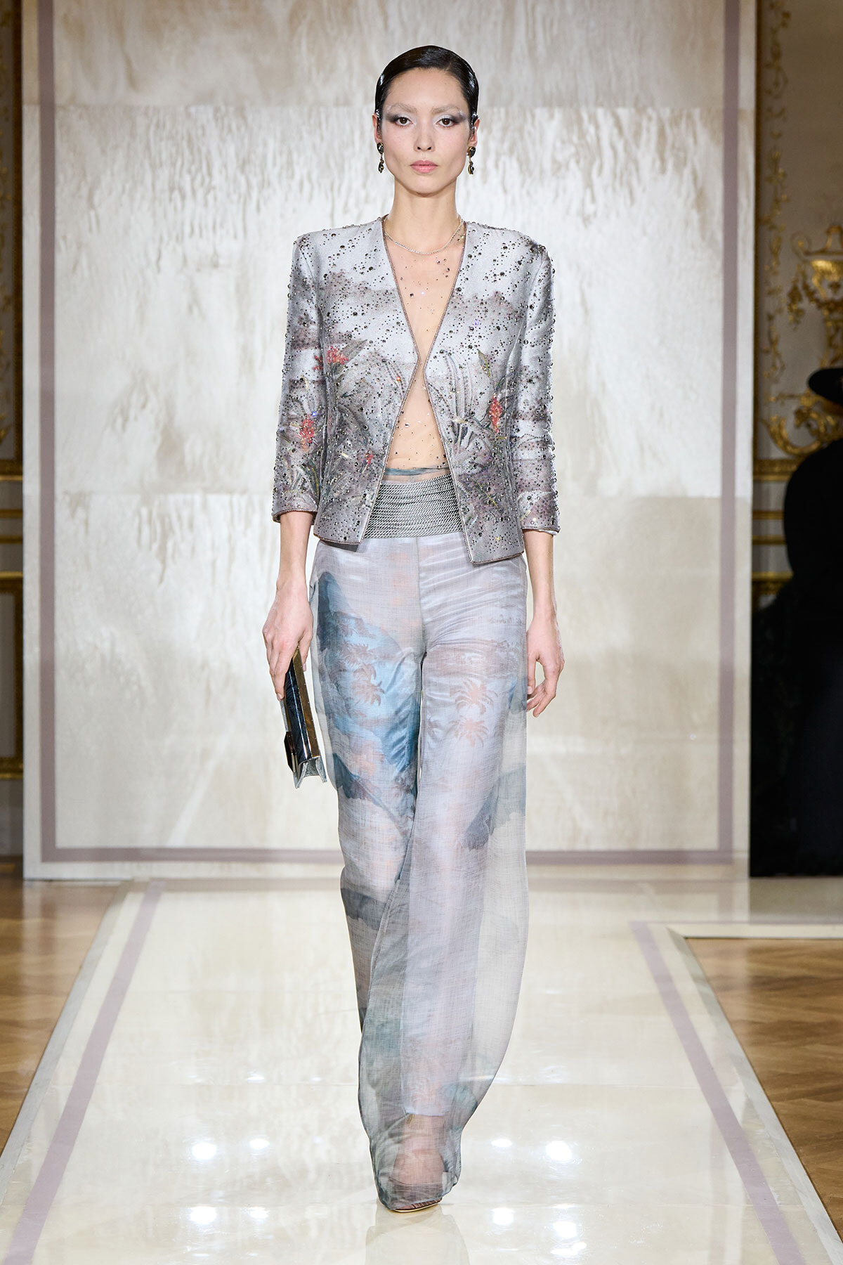 Model on a runway wearing a silver, jeweled blazer with a plunging neckline and sheer, pale-print wide-leg pants, holding a dark clutch.