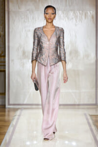 Model on a fashion runway wearing a metallic pink snake-print jacket with a deep V-neck and matching pale pink satin wide-leg pants, holding a small clutch.