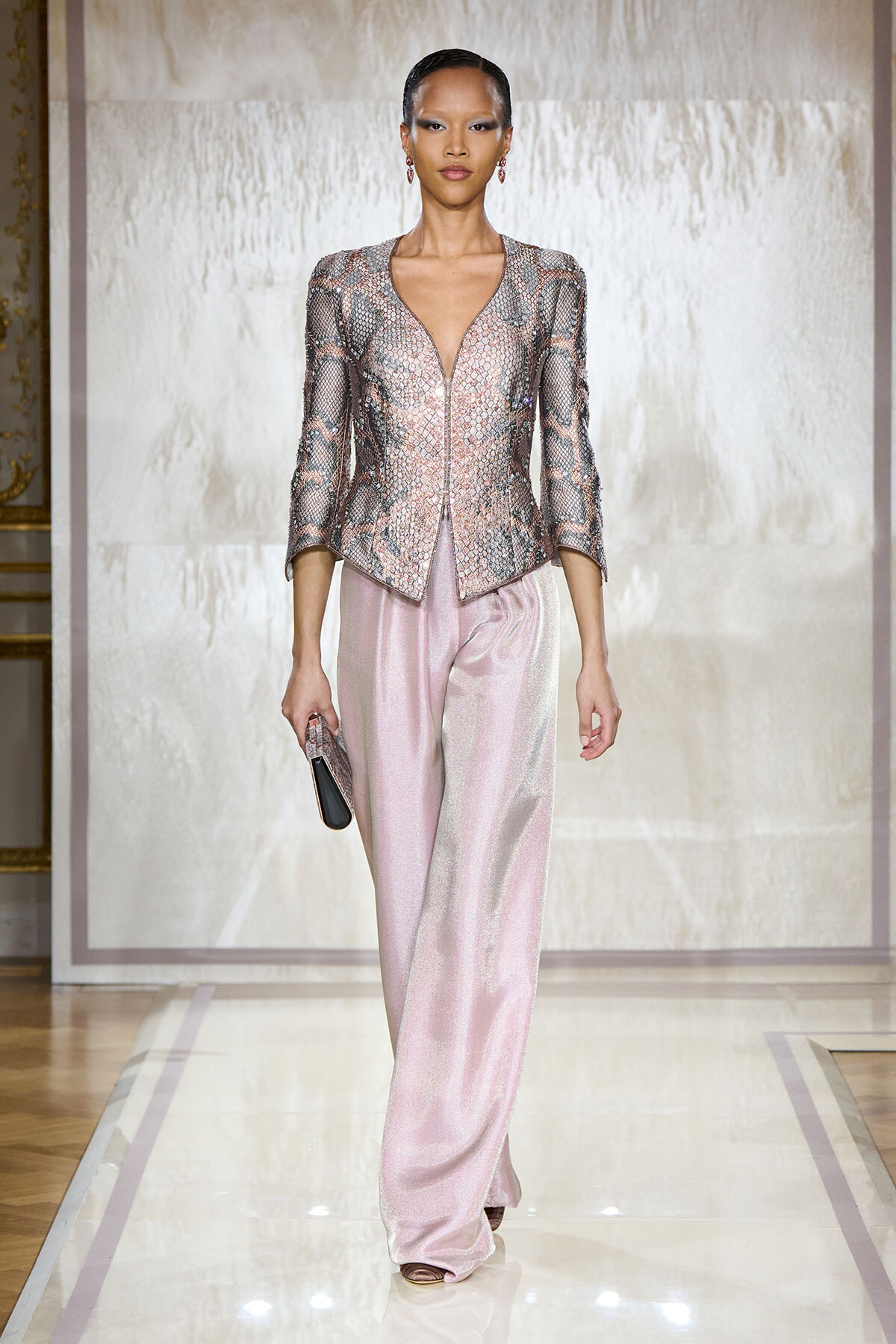 Model on a fashion runway wearing a metallic pink snake-print jacket with a deep V-neck and matching pale pink satin wide-leg pants, holding a small clutch.