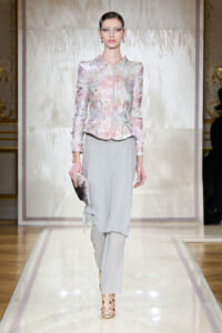 Model on a fashion runway wearing a pastel floral-embroidered jacket with light gray draped trousers, holding a matching clutch.