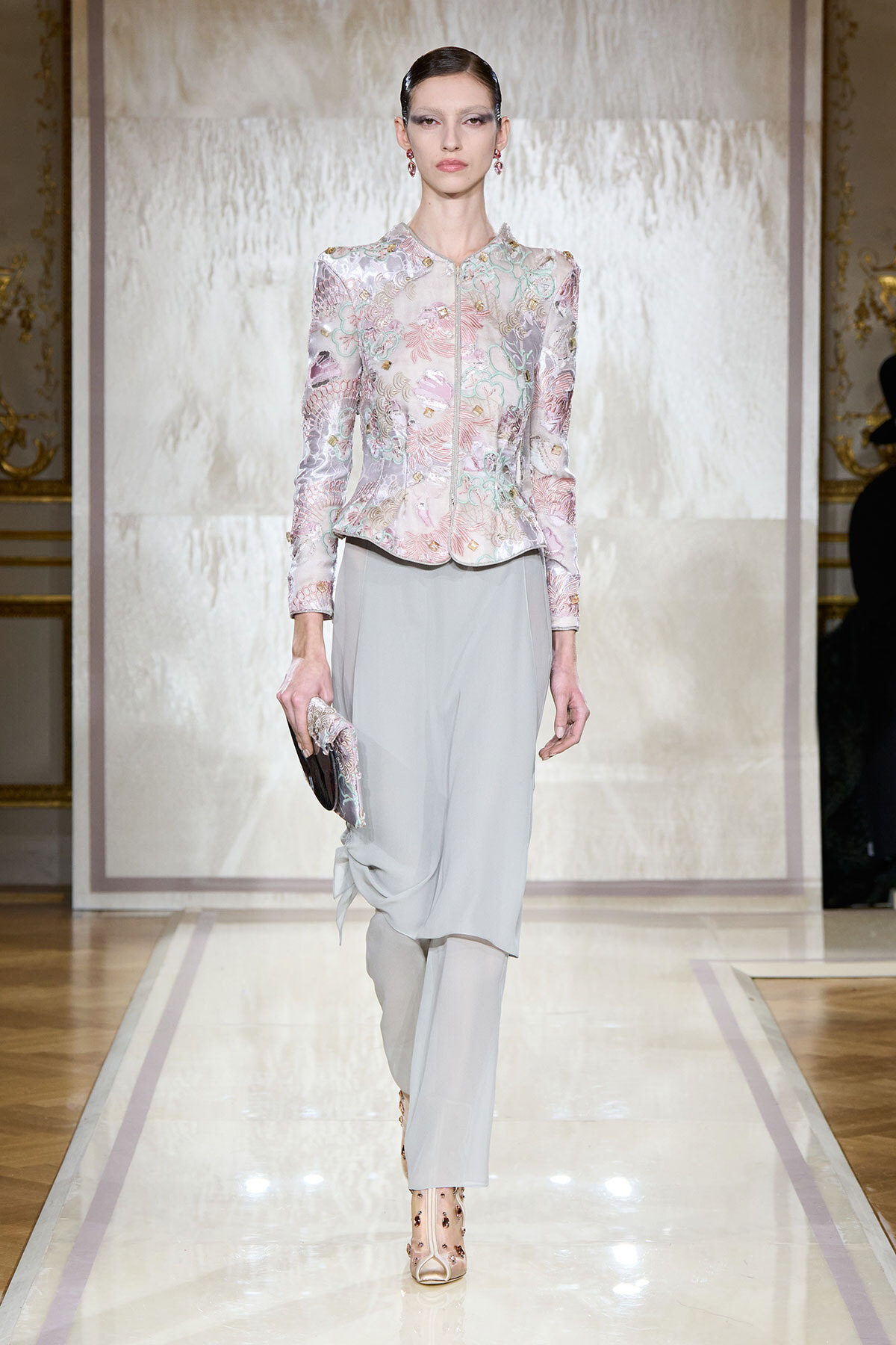 Model on a fashion runway wearing a pastel floral-embroidered jacket with light gray draped trousers, holding a matching clutch.