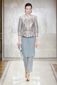 Model on a runway wearing a pastel floral-embroidered jacket over a light blue draped tunic with matching pants, gold ankle boots, and a metallic clutch.