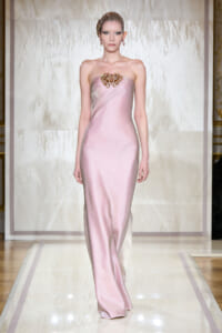 Model walks a pink strapless satin gown with a jeweled chest embellishment on a runway backdrop, wearing a beaded headpiece and earrings.