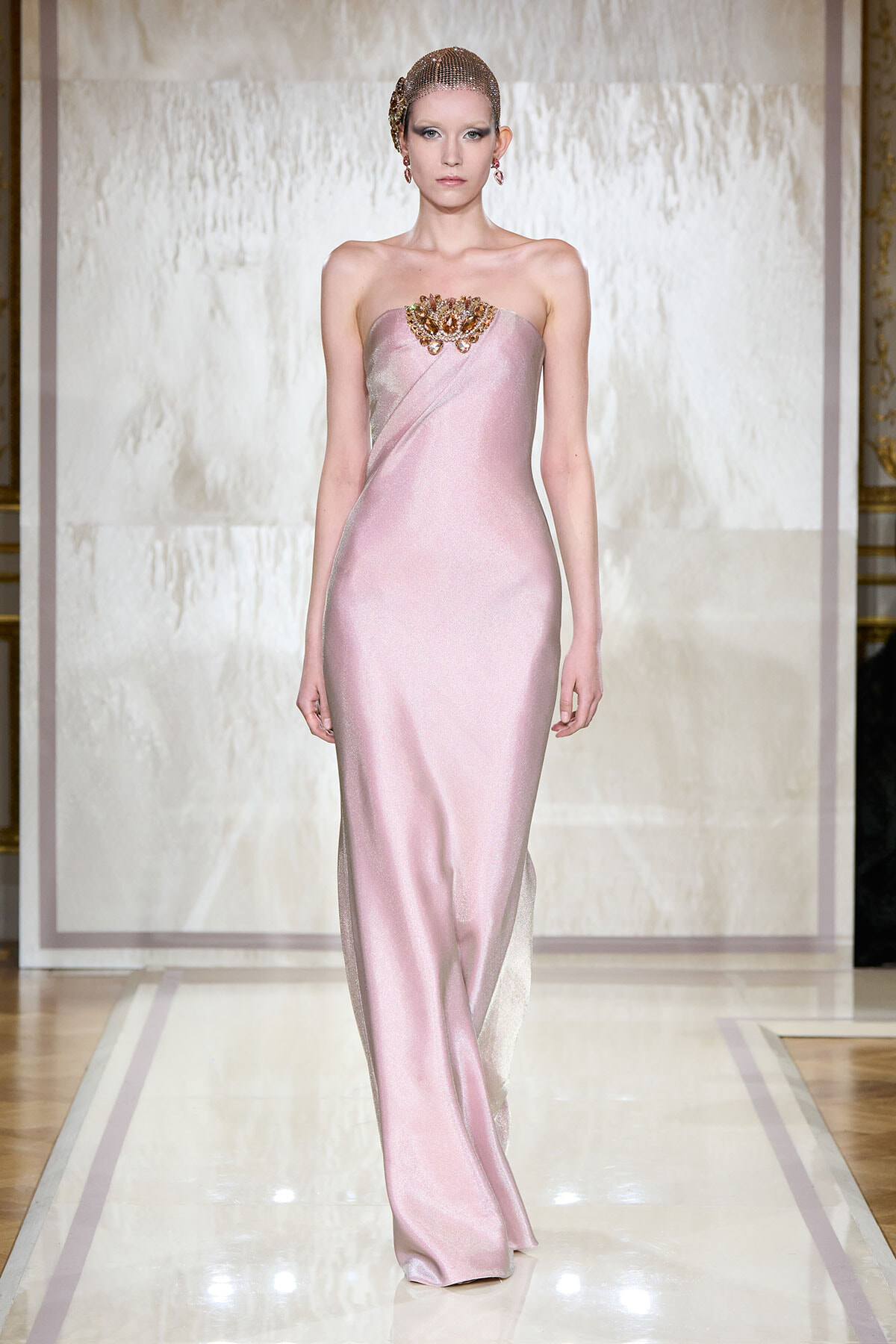 Model walks a pink strapless satin gown with a jeweled chest embellishment on a runway backdrop, wearing a beaded headpiece and earrings.