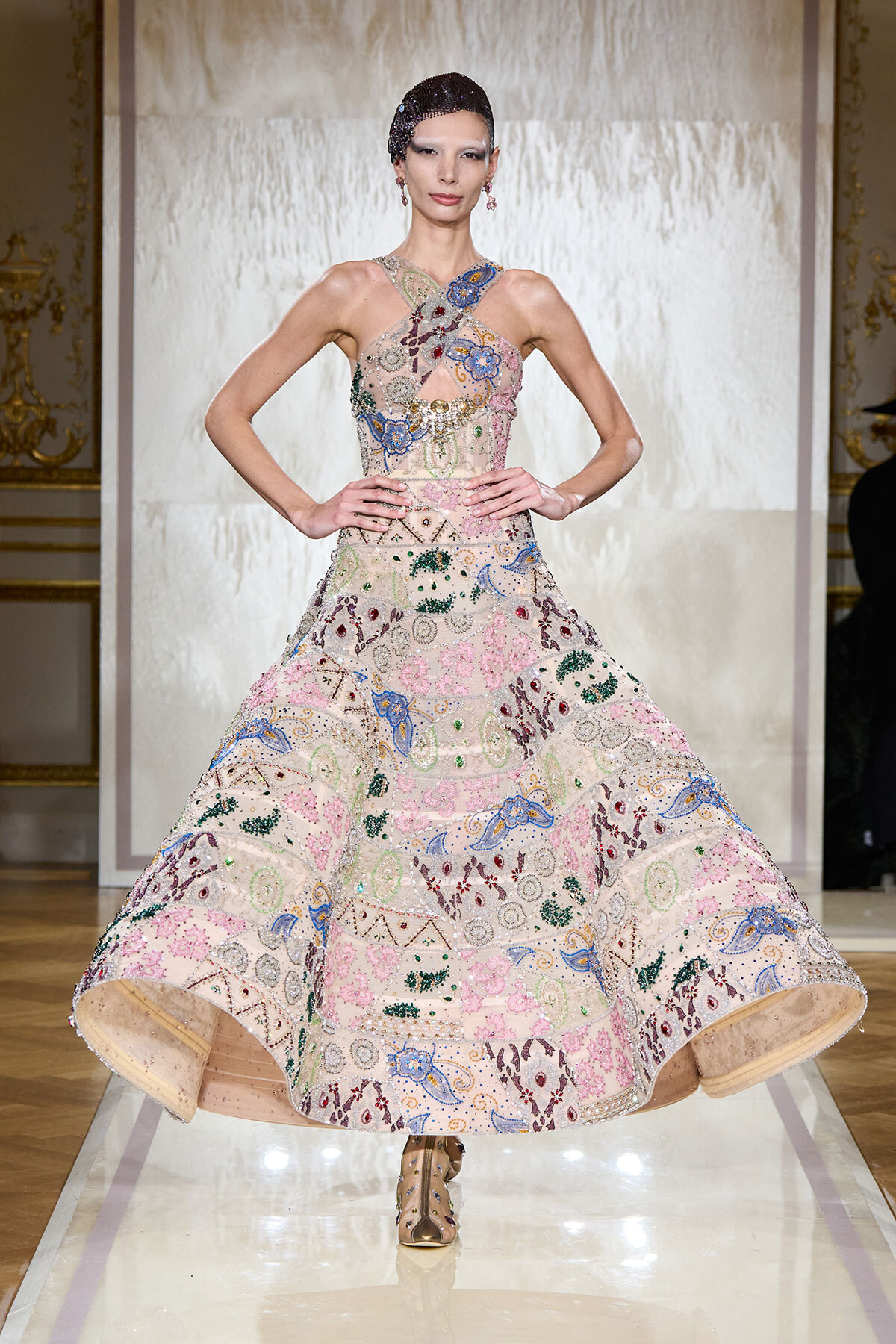 Model on a fashion runway wearing a pastel, heavily embellished floral-embroidered ballgown with a halter neckline, mid-stride.