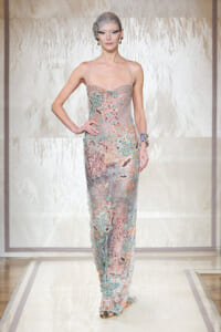 Model on a runway wearing a sheer, pastel-toned gown covered in multicolored embroidery and crystals, with a beaded headpiece.